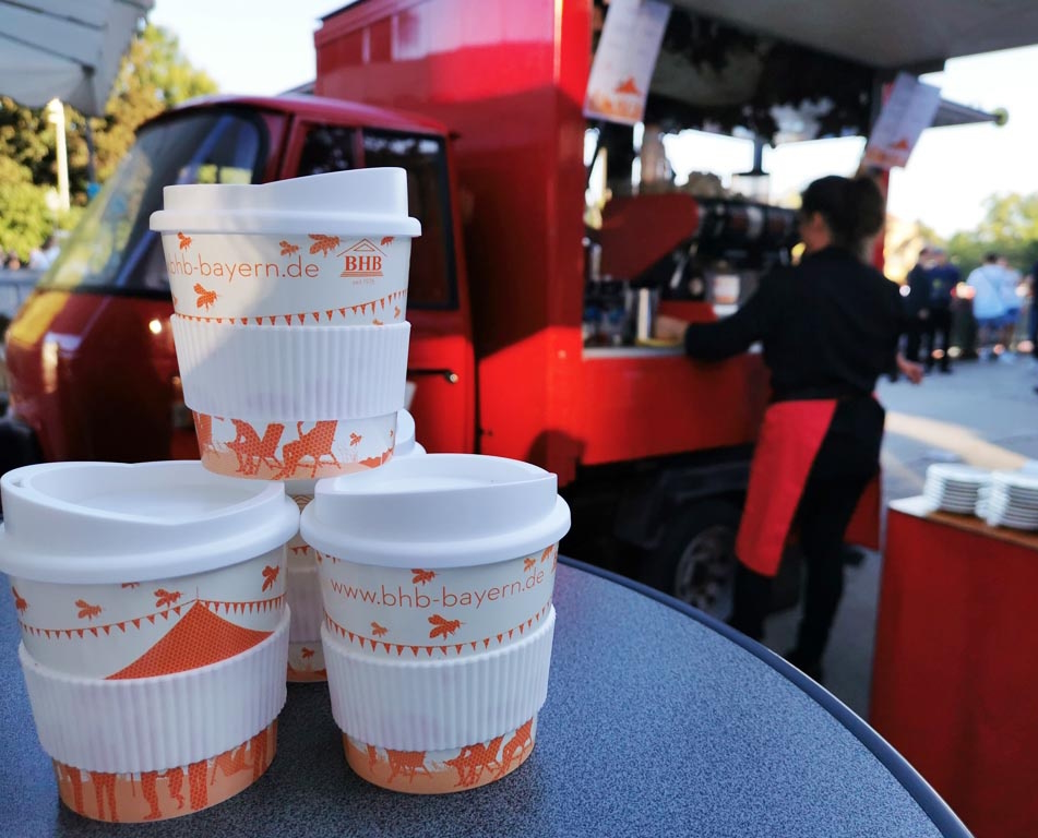 07 Im Hintergrund rotes Espressodreirad, davor gestapelte Mehrweg Kaffeebecher mit Branding und Logo