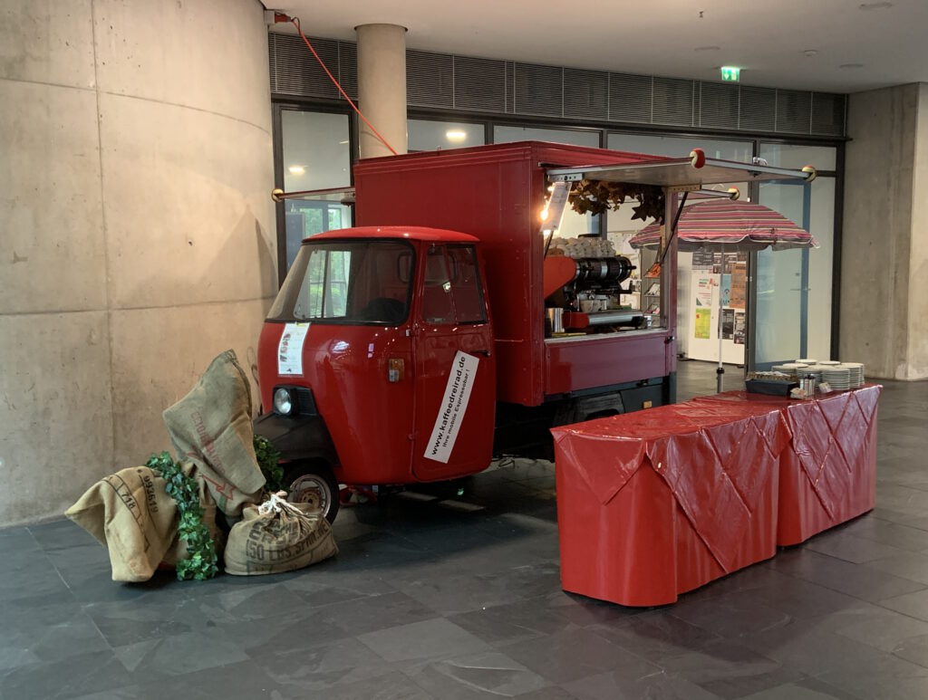 08 Kaffeemobil indoor im Eingangsbereich davor 2 niedrige längliche Tische mit roten Planen eingehüllt, darauf rote Tischdecken